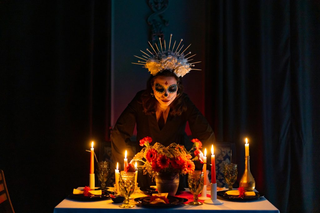 A woman with Day of the Dead makeup stands over a candlelit altar.